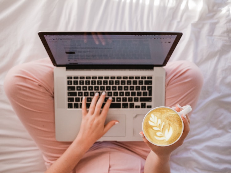 Woman sitting on her bed with laptop and holding a cup of coffee