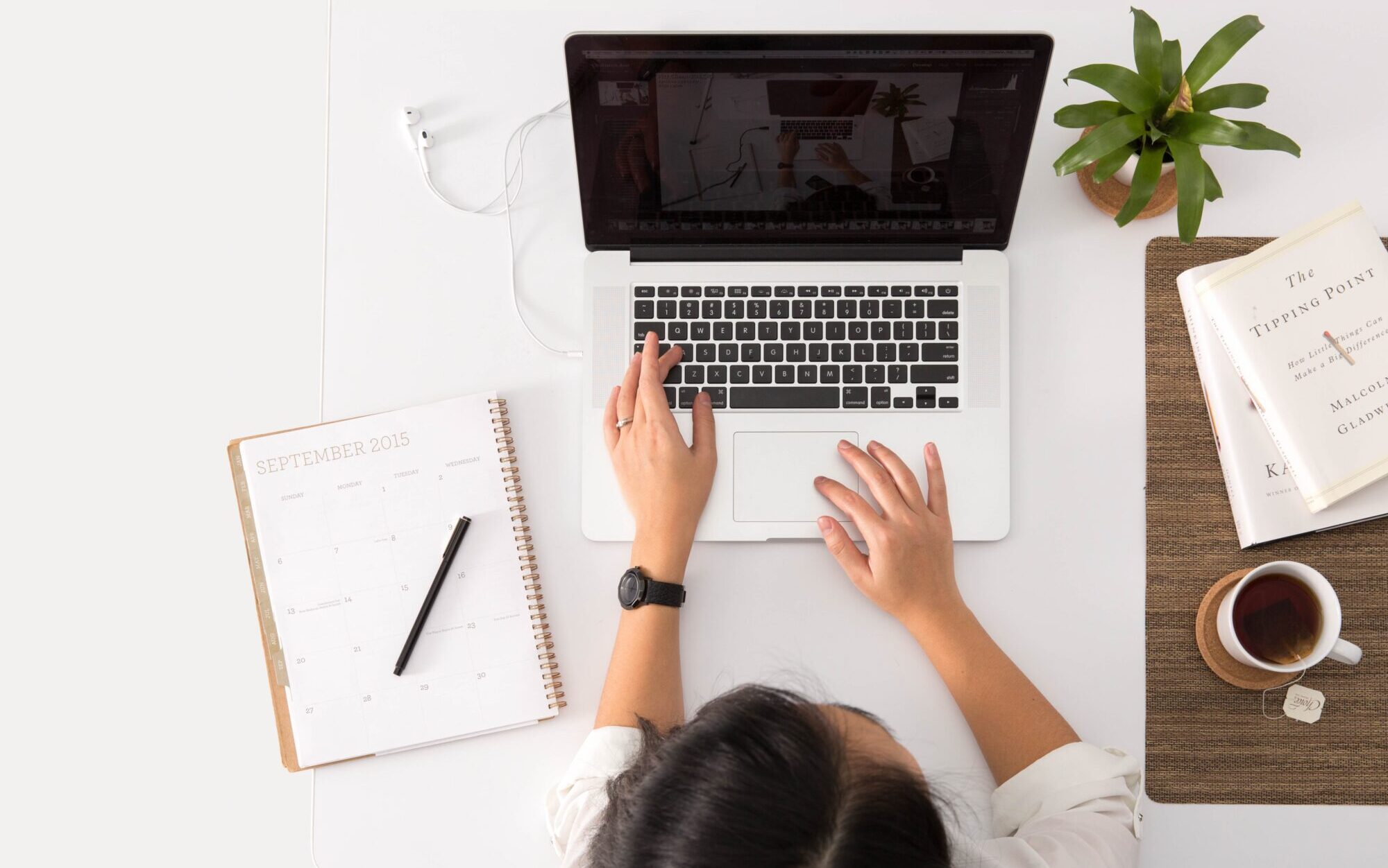 A table with a notebook, coffee cup, plant, a PC and a lady typing on the keyboard. Seen from top to bottom.