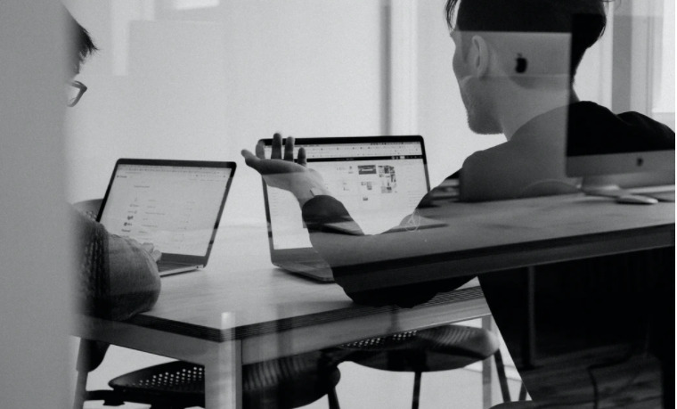 A table with two laptops, and two men discussing something based on the displayed content on their laptops.