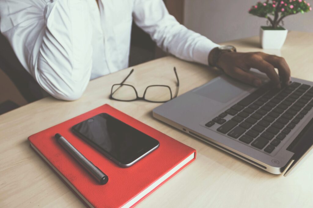 Man working on a laptop placed on a desk. On the desk is a notebook, a small plant, some glasses and a smartphone.