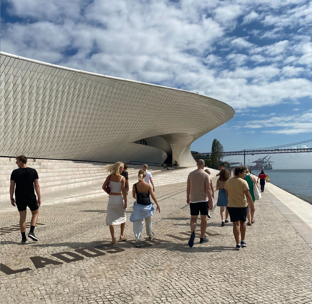 Brostein og flott bygning langs vannkanten i Lisboa, med en gruppe mennesker som går foran. 